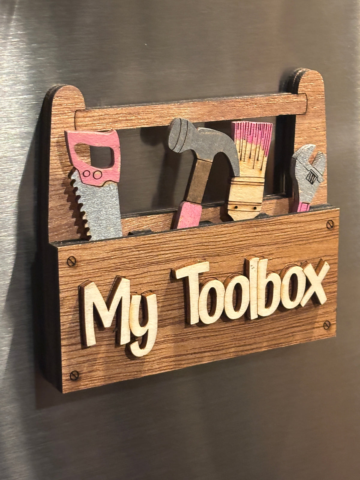 This is a slight side view of the "My Toolbox" . 
This item is shown here on a refrigerator it has 2 Strong Magnets and a Hanger on the back. It also comes with a perfectly sized easel.  
 3d Unique design, Handcrafted, this item is not printed. Its laser engraved and precision cut. 
The tools are removable and can be replaced in any slot you wish.

This item measures approximately 3.5" X 4.0 inches.
