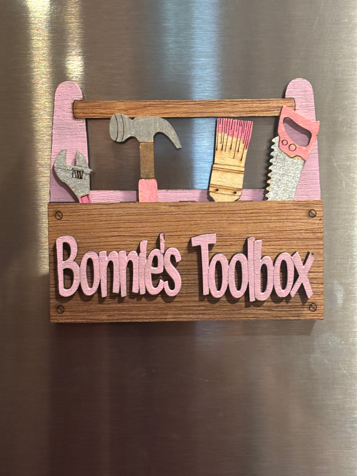 This is a view of a Personalized toolbox.
"Bonnies Toolbox" is shown here on a refrigerator it has 2 Strong Magnets and a Hanger on the back. It also comes with a perfectly sized easel.  
 3d Unique design, Handcrafted, this item is not printed. Its laser engraved and precision cut. 
The tools are removable and can be replaced in any slot you wish.

This item measures approximately 3.5" X 4.0 inches.